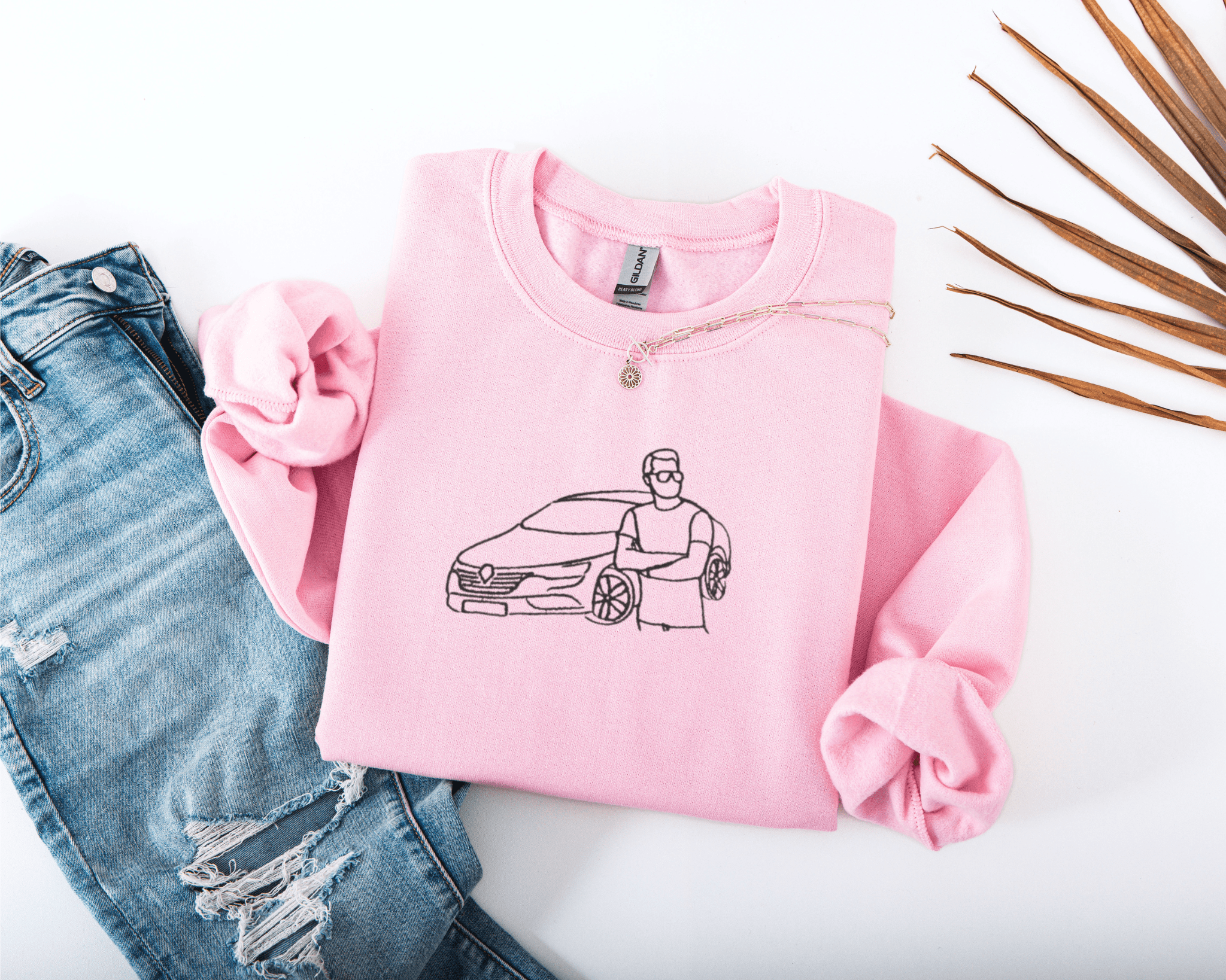 Pink sweatshirt with embroidered design of a car and person, paired with ripped jeans on a white background.