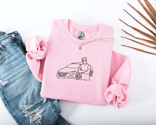 Pink sweatshirt with embroidered design of a car and person, paired with ripped jeans on a white background.